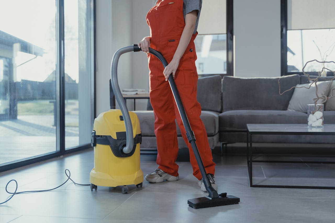 about-03 A professional cleaner in red coveralls vacuuming a modern living room with a yellow vacuum cleaner.